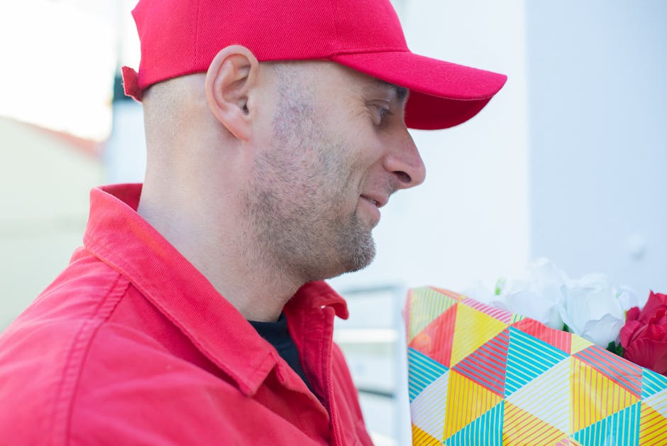 A delivery person wearing a red cap and matching red jacket is holding a colourful gift bag filled with fresh flowers, including white and red blooms. The bouquet features vibrant petals and lush gree