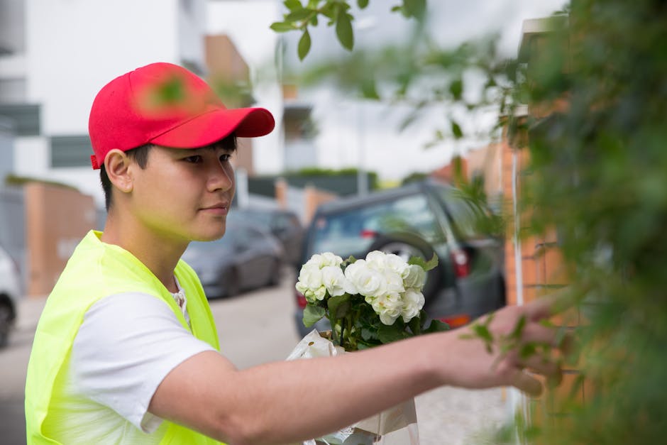 A young delivery person wearing a red cap and a bright yellow high-visibility vest is shown outdoors, holding a bouquet of fresh white flowers with green foliage. The bouquet is wrapped in white paper