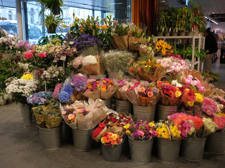 A large display of fresh flowers arranged in metal buckets and wrapping paper, featuring a variety of colourful blooms including pink roses, purple lilacs, white chrysanthemums, yellow sunflowers, and