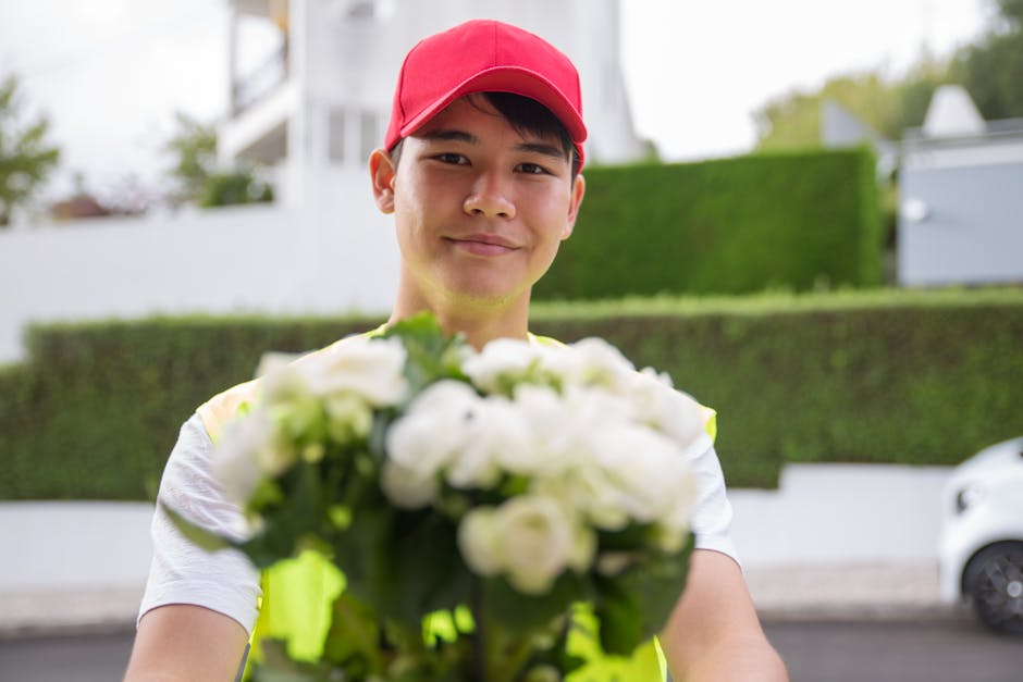 A delivery person wearing a red cap and a high-visibility vest extends a bouquet of fresh white roses, peonies, and greenery towards the camera. The arrangement is wrapped in clear cellophane and tied