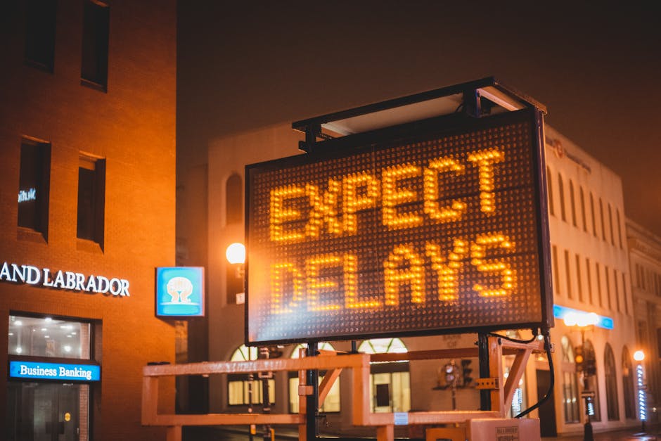 A digitally illuminated outdoor LED sign displaying the message 'EXPECT DELAYS' in bright orange text against a dark evening sky. The sign is mounted on a metal frame on a city street, with nearby bui